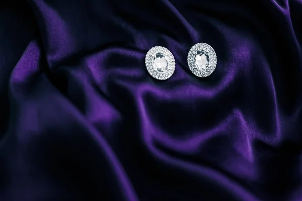 Luxury diamond earrings on dark violet silk fabric, holiday glam