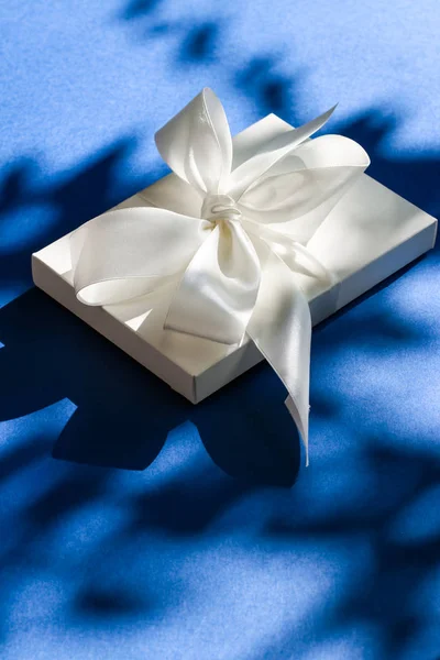 Luxury holiday white gift box with silk ribbon and bow on blue b