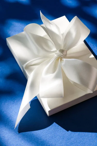 Luxury holiday white gift box with silk ribbon and bow on blue b
