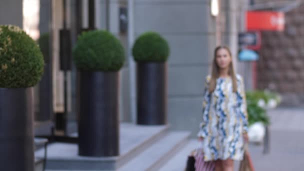 Giovane bella ragazza sta camminando lungo la strada della città con le borse della spesa dopo uno shopping di successo. Bruna in un vestito. Rallentatore 4k — Video Stock