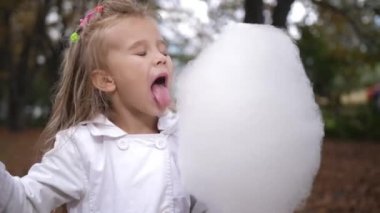 Küçük sarışın kız şehir parkında tatlı pamuk şeker yiyor. Helva yemek güzel küçük bir kız. Pamuk şeker yeşil orman backgorund, yeme çocuk. 4 k. ağır çekim