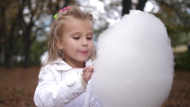 Маленькая блондинка ест сладкую сахарную вату в городском парке. Красивая маленькая девочка ест сладкую нитку. Ребенок ест сахарную вату зеленый лес на backgorund. 4k. Slow Motion — стоковое видео