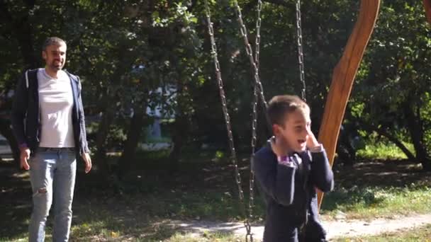 Feliz joven padre y sus hijos jugando en el columpio en el parque. Feliz padre sonriente enrolla a su hijo en un columpio en el parque. 4k. Movimiento lento — Vídeo de stock