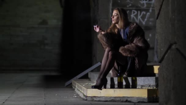 Plan général. Sexy girl dans un manteau de fourrure assis sur les marches fume et boit du vin. Ivre femme seule dans une ville de nuit 4K Slow Mo — Video