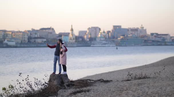 Пара делает селфи на пляже осенью на фоне реки и города. 4K Slow Mo — стоковое видео