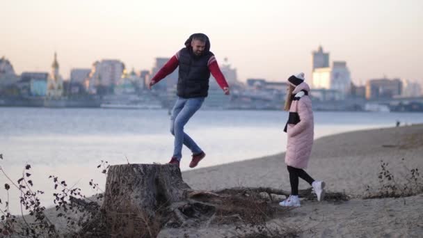 Счастливая молодая пара, гуляющая по пляжу осенью на фоне реки, становясь на пень улыбаясь и обнимая 4K Медленно Мо — стоковое видео