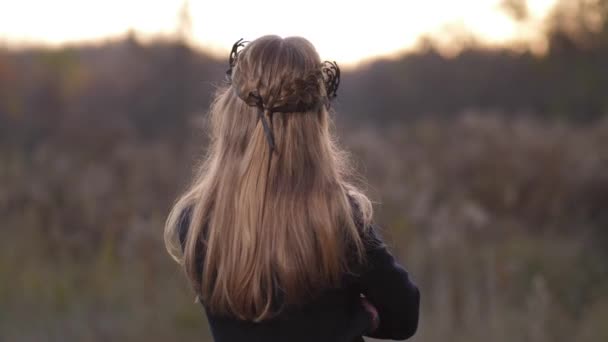 De cerca, una chica con el pelo rubio está de pie con la espalda y luego se gira con una expresión seria en su cara. Lleva una máscara y un abrigo. Otoño, campo, puesta del sol 4K Lento Mo — Vídeo de stock