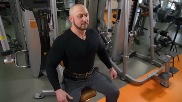 Adult bodybuilder sits on the simulator and spreads his arms around, lifting a lot of weight 4K Slow Mo — Stock Video