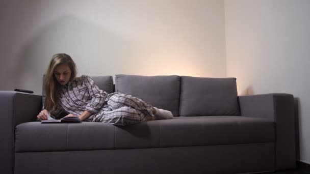 Una chica acostada en el sofá leyendo un libro, ella está en ropa de casa. Pasa tiempo libre pacíficamente. 4K Slow Mo — Vídeo de stock