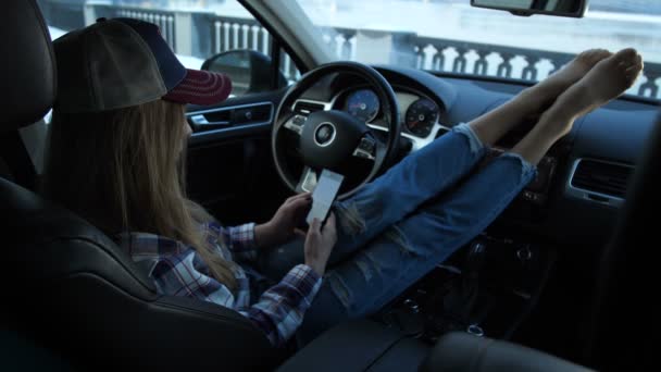 Flickan sitter i bilen. Sitt ben lie på panelen, flickan ser ut in i telefonen, för ram av vintern. 4 k långsamma Mo — Stockvideo