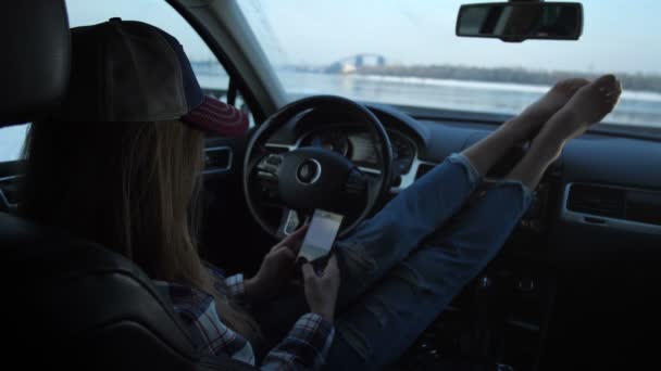 Dziewczyna siedzi w samochodzie. Ona pisze Sms. zewnątrz okna jest piękny krajobraz, rzeki i most. 4 k Slow Mo — Wideo stockowe
