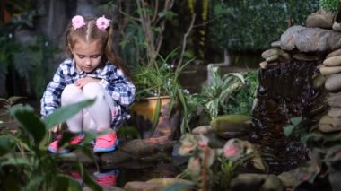 Güzel tropik bahçe. Suyun kenarında küçük bir kız oturuyor ve parmağını suyla dokunur. 4 k yavaş Mo