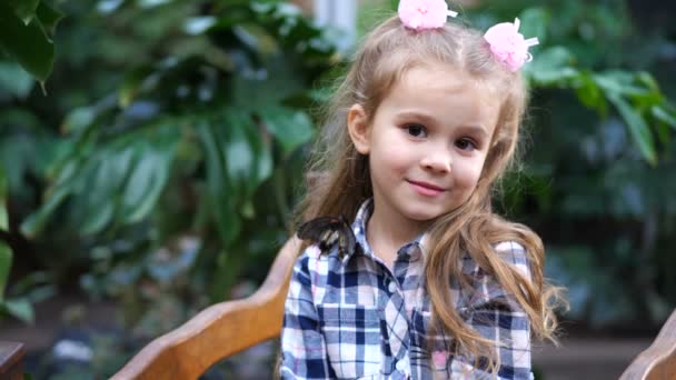 La fille avec un sourire sur le visage regarde dans la caméra. Elle est assise sur le pont dans le jardin, sur son épaule un papillon. Mo lent 4K — Video