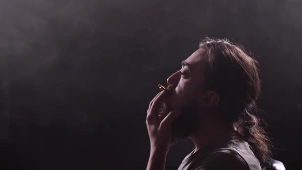 Un type avec une longue barbe et oblique fume une cigarette sur un fond noir. Libère des anneaux de fumée. Mo lent 4K — Video