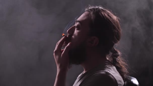 Um homem com barba e cabelo comprido fuma um cardume num quarto escuro, muito fumo à sua volta. 4K Mo lento — Vídeo de Stock