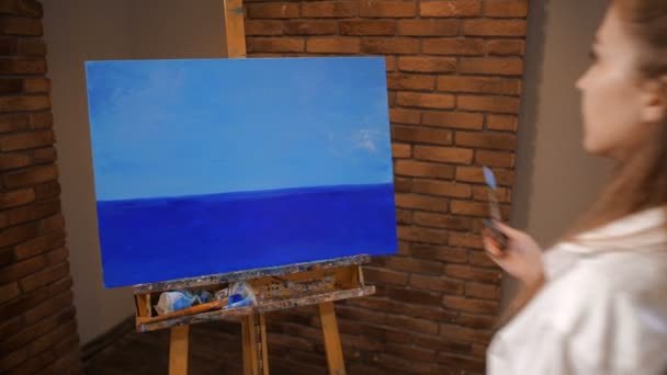 Obraz jest na sztalara, Dziewczyna pojawia się i zaczyna rysować. Maluje krajobraz, niebo i morze. 4K slow mo — Wideo stockowe