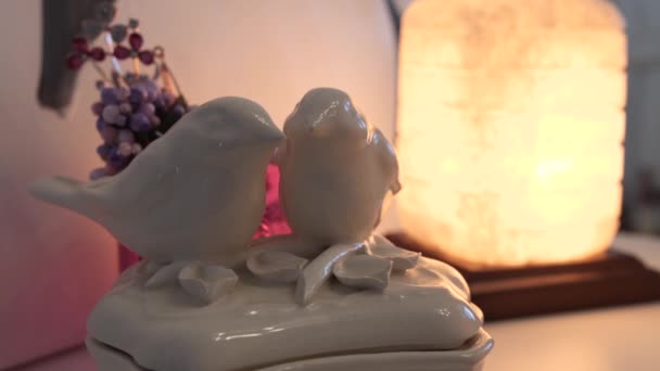 Figurinha de porcelana na forma de dois pássaros amorosos em pé na prateleira contra o fundo da lâmpada no interior da casa. 4K Mo lento — Vídeo de Stock