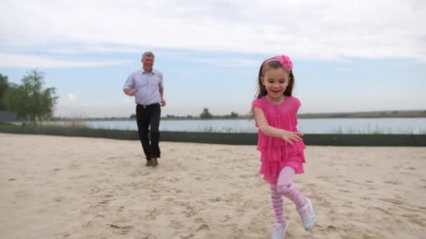 En liten, vacker flicka som kör med sin farfar på stranden. Flickan körs på förgrunden, farfar i Defocus på bak. 4K långsam Mo — Stockvideo