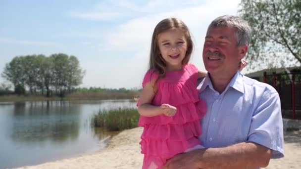 Grand-père et petite-fille marchent le long de la plage. La fille est assise avec son grand-père sur ses mains. Ils s'amusent à rire. Mo lent 4K — Video