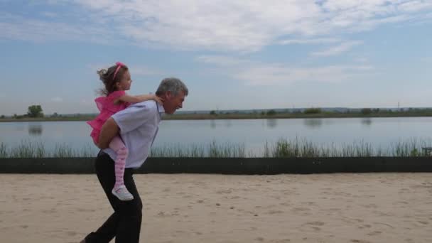 Kameran är i rörelse. Flickan går längs stranden med hennes barnbarn på ryggen. Liten flicka jublar. 4K långsam Mo — Stockvideo