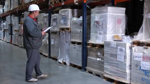 El trabajador del almacén reescribe la mercancía. Un hombre con un casco y un uniforme con una carpeta llegando a la caja. 4K Slow Mo — Vídeos de Stock