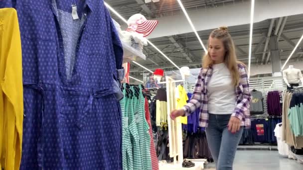 Grote winkel. Het meisje loopt tussen de rijen met kleren, neemt een T-shirt en bladeren. 4k slow mo — Stockvideo