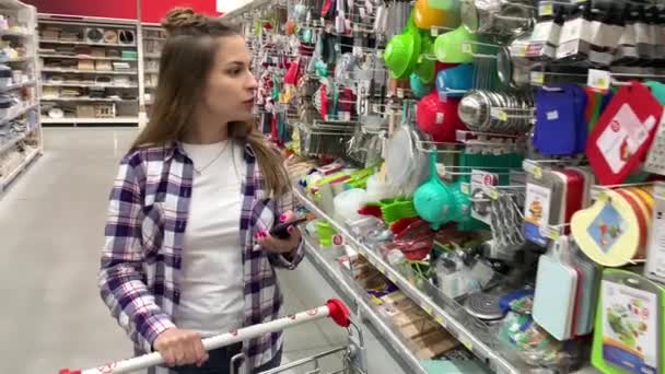 Молодая домохозяйка ходит с тележкой вокруг супермаркета, смотрит на список покупок по телефону. 4K Slow Mo — стоковое видео