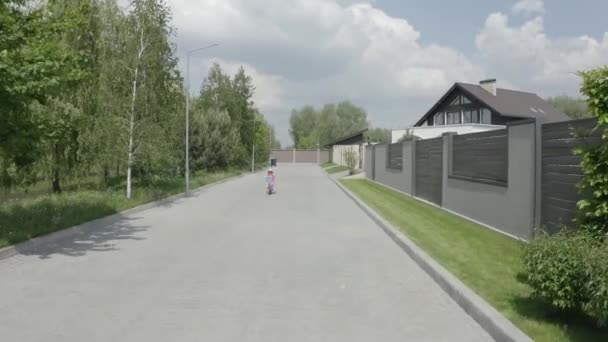 Стрельба с беспилотника. Маленькая девочка едет по велосипедной дорожке вдоль части домов. 4K Slow Mo — стоковое видео