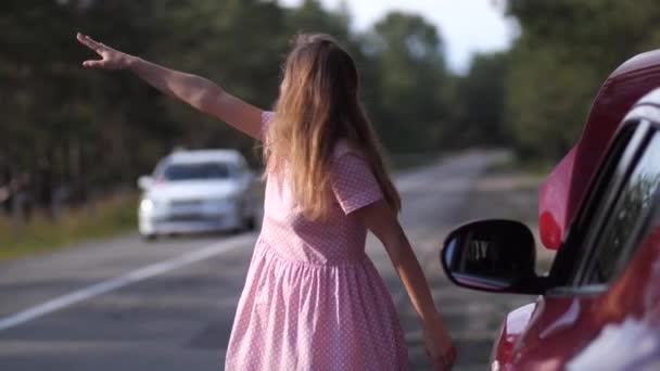 Блондинка стоит у дороги рядом со сломанной машиной. Девушка пытается остановить кого-то за помощью. 4K Slow Mo — стоковое видео