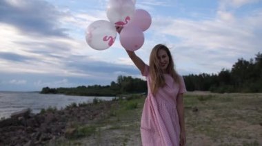 Pembe elbiseli genç bir sarışın sahilde toplarla yürüyor. Sonra da topları kameraya veriyor. 4k Ağır çekim