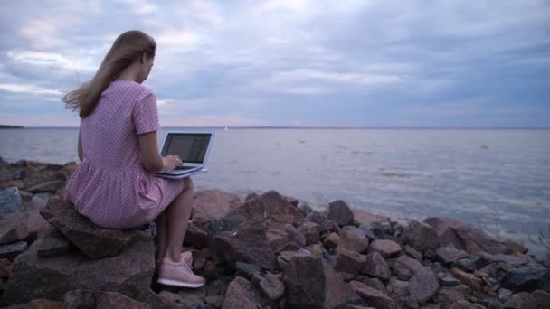 A szőke a köveken ül a tenger mellett. Egy laptopon gépel. Inspirációt keresek. 4k Lassú Mo — Stock videók