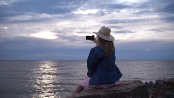 Una mujer joven se sienta en una piedra y toma una foto por teléfono. La chica fotografía el cielo del atardecer. 4K Slow Mo — Vídeos de Stock