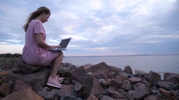 Freelancer trabalha fora do escritório. Uma rapariga junto ao mar a trabalhar num portátil. 4K Mo lento — Vídeo de Stock