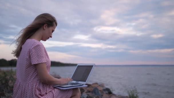 Девушка работает на ноутбуке на пляже у моря. Ветер красиво дует ей на волосы. 4K Slow Mo — стоковое видео