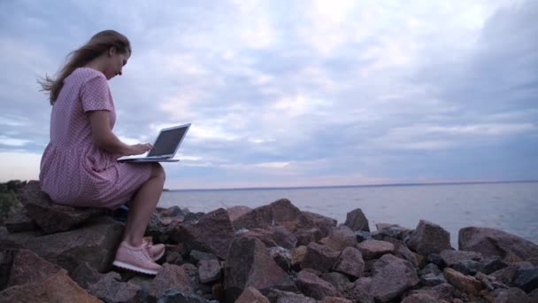 Работать на улице. Женщина на пирсе у моря печатает текст на ноутбуке. 4K Slow Mo — стоковое видео