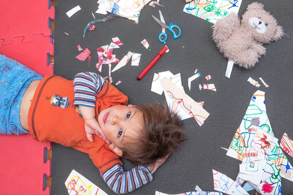 Creative chaos. A little boy is lying on the floor among the scattered and chopped drawings.