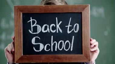 schoolboy covering face with blackboard with back to school lettering and smiling while looking at camera 