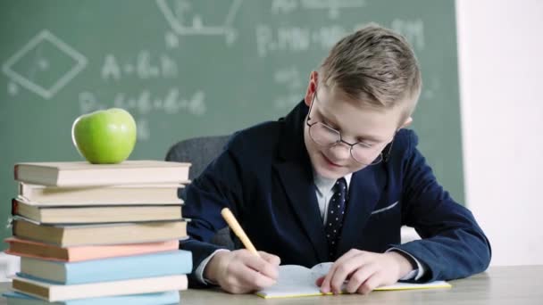 šťastný školák v brýlích, který píše v poznámkovém bloku u knih a jablek a ve škole ukazuje palce 