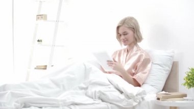 dijital tablet kullanarak çekici sarışın genç kadın ve yatakta otururken gülümseyen 