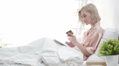 kredi kartı bakarak çekici sarışın kız, dijital tablet kullanarak ve yatakta otururken gülümseyen 