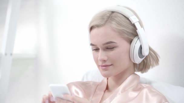 Happy Blonde Girl Touching Headphones While Listening Music Using Smartphone — Stock Video