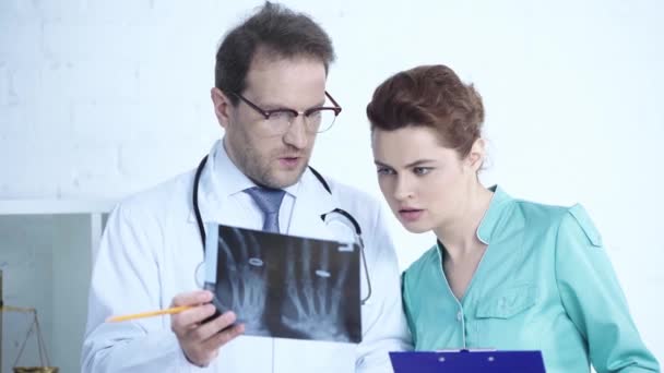深刻な医師と一緒にX線診断を見て クリップボードに書き込むかわいい看護師 — ストック動画