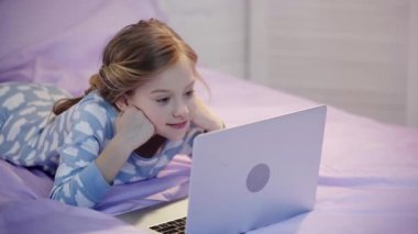 pijama içinde preteen çocuk yatakta yatan, el ile yüz propping ve gülümseme ile dizüstü bilgisayar kullanarak