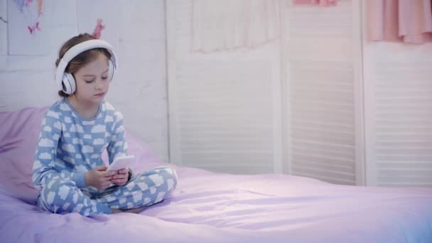 穿着睡衣的孩子坐在床上 在耳机里听音乐 使用智能手机 — 图库视频影像
