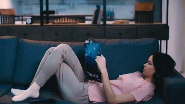 自宅でソファに横たわって科学雑誌を読むかわいい若い女性 — ストック動画