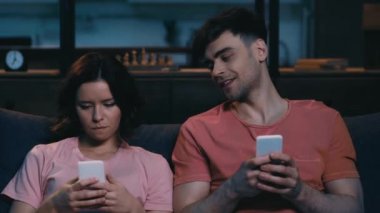 neşeli erkek ve kadın akıllı telefonlar kullanarak, gülümseyerek ve evde kanepede otururken birbirlerine bakıyor