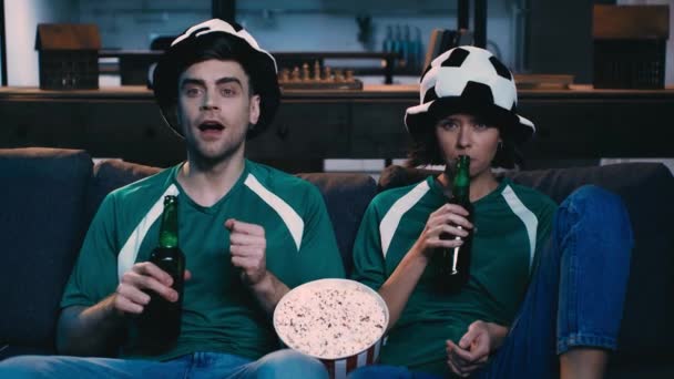 自宅でテレビでサッカーの試合を見ながらビールを飲んだり ポップコーンを食べたりするファン帽子に集中した男女 — ストック動画