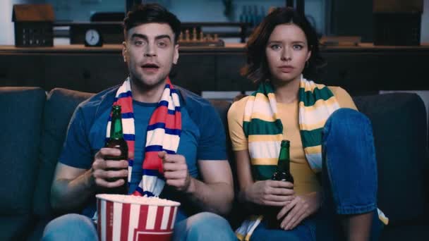 自宅のソファで動揺した男の近くでサッカーの試合を見ながら勝者のジェスチャーを示す興奮した若い女性 — ストック動画