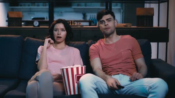 退屈な若い男女がソファに座って ポップコーンを食べ テレビを見ながらリモコンを使う — ストック動画