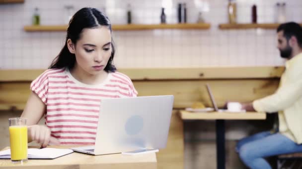 ノートに書き込み ラップトップに入力するTシャツのアジアの女性 コンピュータを使用して バックグラウンドでコーヒーを飲む男性 — ストック動画